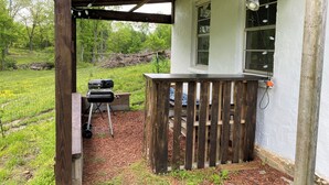 A covered outdoor area with a grill, a wooden deck, and a window overlooking a grassy yard. - Tranquil 2-bedroom house with AC, fire pit, & game room in the Ozarks (Marshall)
