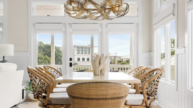 A dining area with a round table and four wicker chairs, a hanging light fixture, and large windows with a view of buildings outside. - Private Pool | Great for Families | Tides of Joy (Isle of Palms)