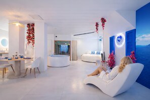 A modern living room with a white sofa, a dining area with a round table, and a view of the sea through a large window. - Love Room Pézenas – Private Hot Tub & Sauna – Suite Santorini (Pézenas)