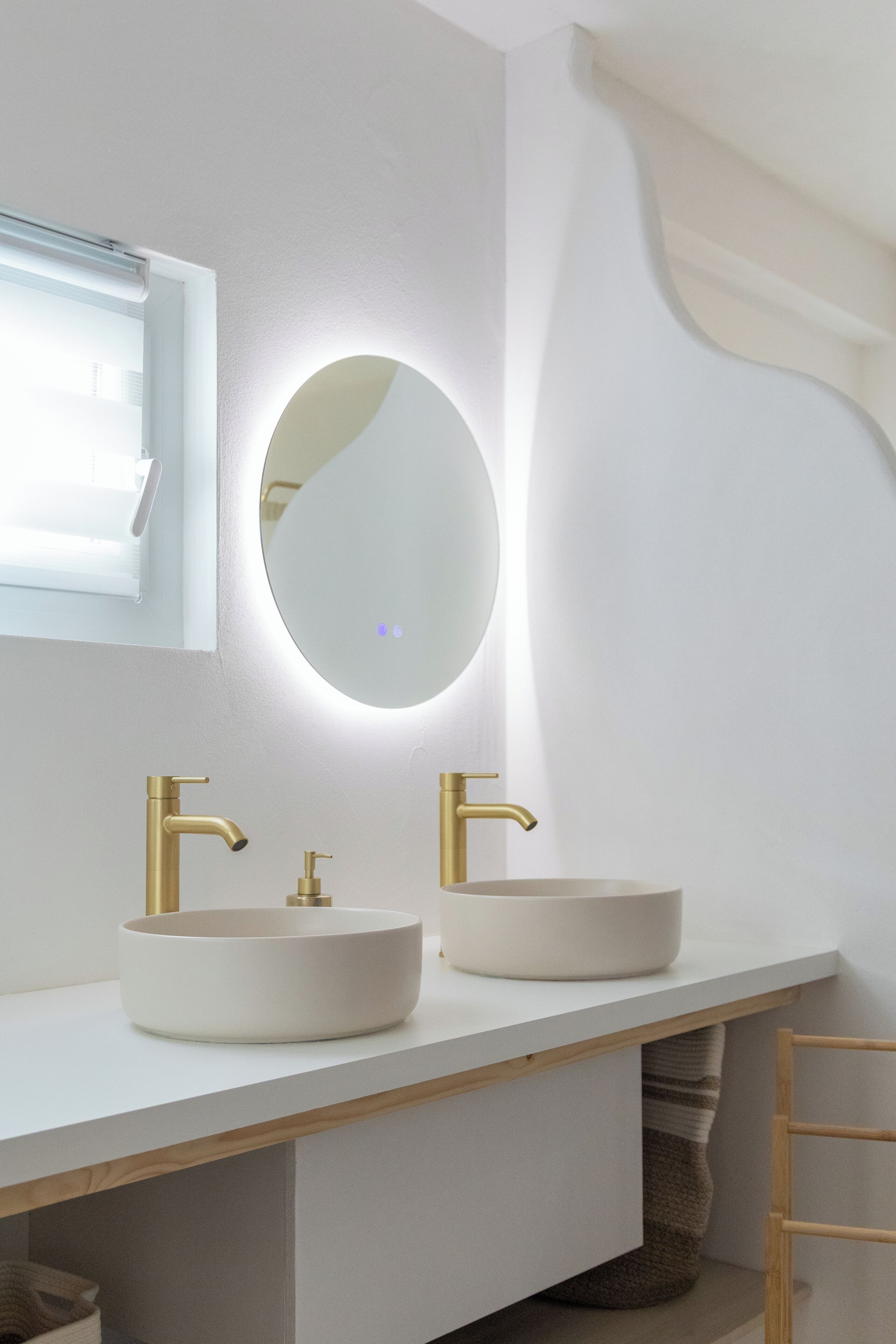 Un baño moderno con dos lavamanos blancos, grifos dorados y un espejo redondo con luz.