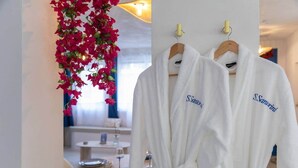 Et hotelværelse med hvide badekåber på en træstativ, et rundt bord med et glas vin og en blomsterophæng.