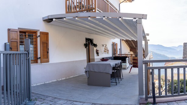 A covered outdoor dining area with a table and chairs, a balcony with wooden railings, and a view of mountains in the background. - Casa Del Torchio: Apartment with vineyard view in Trentino (Valternigo)