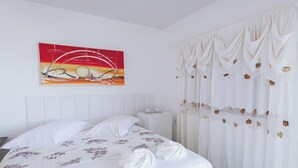 Una cama bien tendida con sábanas blancas y un estampado floral, una mesita de noche blanca con una taza y un platillo, y un cuadro colgado en la pared que representa un atardecer.
