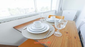 Een eettafel gedekt met witte borden, een kop, een wijnglas en een vork, met uitzicht op het buitengebeuren door een raam.
