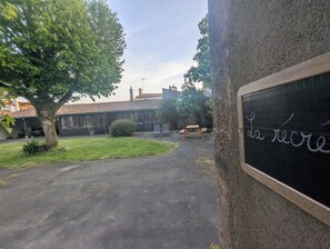 Une cour avec un banc en bois, un tableau noir sur lequel est écrit « da recrea », et un bâtiment avec un toit en tuiles.