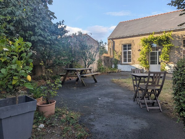 Une cour avec deux tables et des chaises en bois, entourée de plantes en pot et bordée d’un bâtiment dont le toit est recouvert de tuiles.