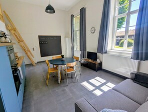 Ein Zimmer mit Esstisch, Stühlen, einer Couch, einem Fernseher, einer Tafel und einem Fenster mit Vorhängen.