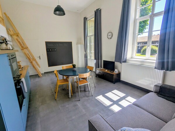 Ein Zimmer mit Esstisch, Stühlen, einer Couch, einem Fernseher, einer Tafel und einem Fenster mit Vorhängen.