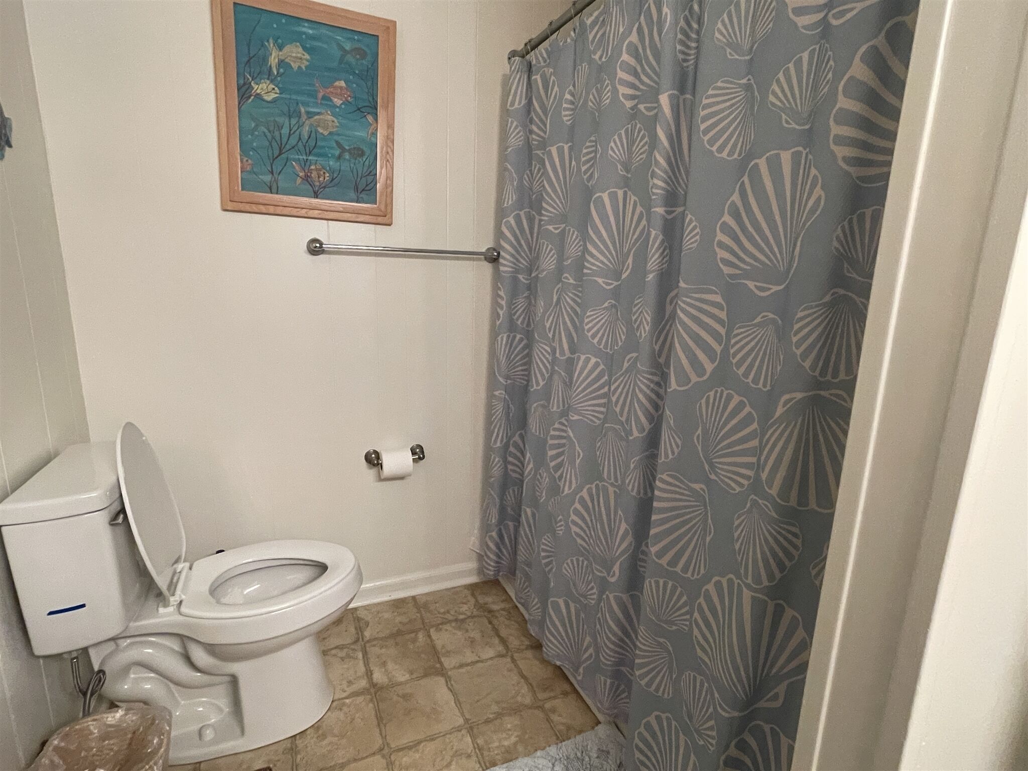 Une salle de bain équipée de toilettes, d’un porte-serviettes, d’un rideau de douche orné de motifs de coquillages et d’un tableau encadré accroché au mur.