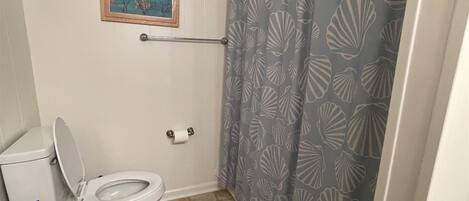 Une salle de bain équipée de toilettes, d’un porte-serviettes, d’un rideau de douche orné de motifs de coquillages et d’un tableau encadré accroché au mur.
