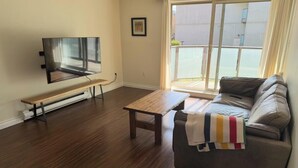 A living room with a sofa, a coffee table, a bench, and a TV. - 2 bedroom condo available for FIFA (Burnaby)