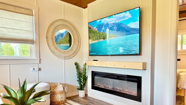 A living room with a TV mounted on the wall, a fireplace below, and a window with blinds. - Anchor Cabin | Walk to Lake Como + Fire Pit + Grill (Lake Geneva)