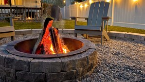 A stone fire pit with logs, surrounded by gravel, wooden chairs, and a white fence with string lights. - Anchor Cabin | Walk to Lake Como + Fire Pit + Grill (Lake Geneva)