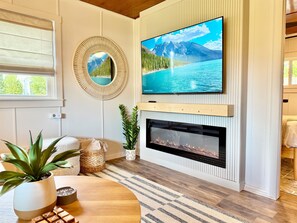 A living room with a TV mounted on the wall, a fireplace below, and a window with blinds. - Anchor & Oak Cabins | Lake Como Retreat – 2 Cabins, 4 Bed/2 Bath - Sleeps 7 (Lake Geneva)