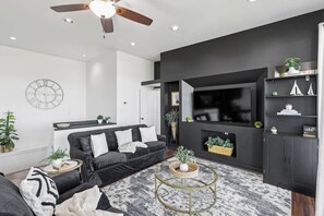 A modern living room with a black sofa, a round gold coffee table, and a large flat-screen TV mounted on the wall. - Gated Community Get Away (Frisco)