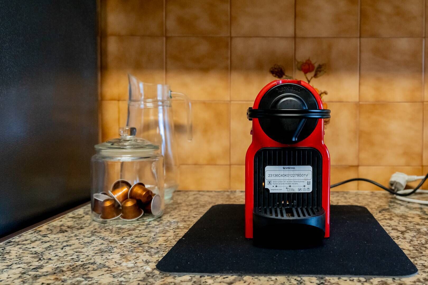 Una máquina de café Nespresso roja sobre una encimera de cocina, junto a un tarro de vidrio con cápsulas de café.