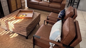 Un vaste espace de vie comprenant un canapé en cuir marron, une table basse en verre et un pouf en osier. Au fond, on aperçoit un coin cuisine avec une table à manger et des chaises.
