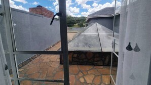 Vista através de uma porta de vidro para um pátio com piso de pedra, grade de metal, parede de tijolos e um edifício cinza com telhado inclinado.