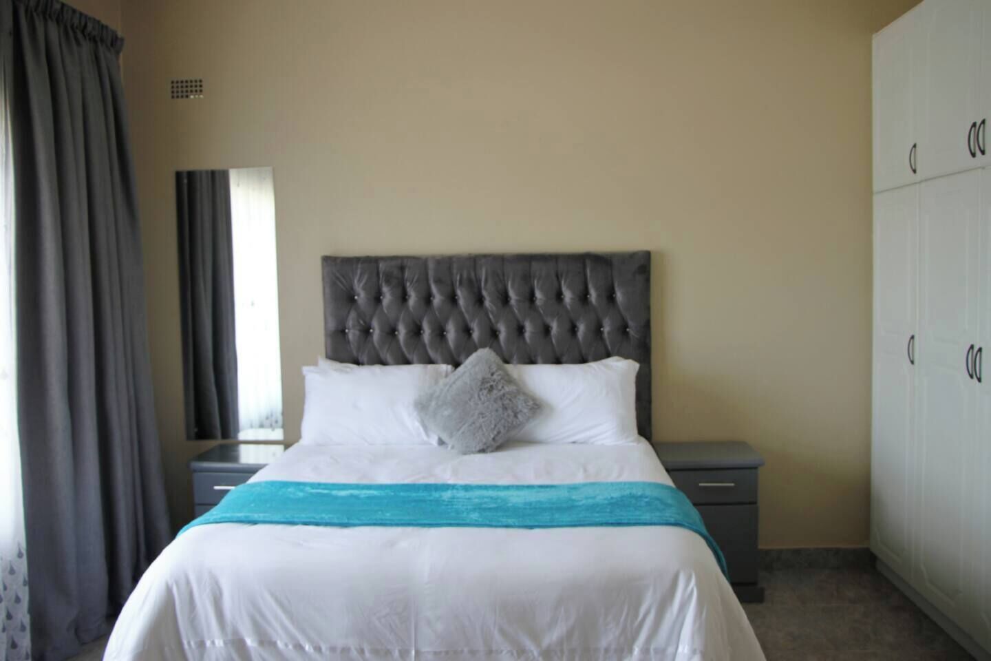 A bedroom with a tufted headboard, a bed with white bedding and a teal throw, a nightstand, a wardrobe, and a window with curtains.