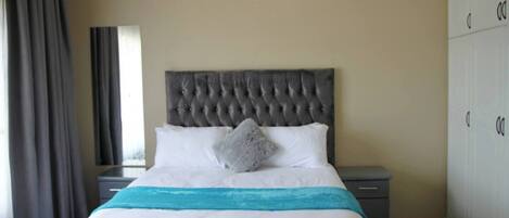 A bedroom with a tufted headboard, a bed with white bedding and a teal throw, a nightstand, a wardrobe, and a window with curtains.