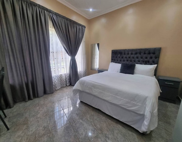 A bedroom with a large bed, a nightstand, a chair, and a window with curtains.