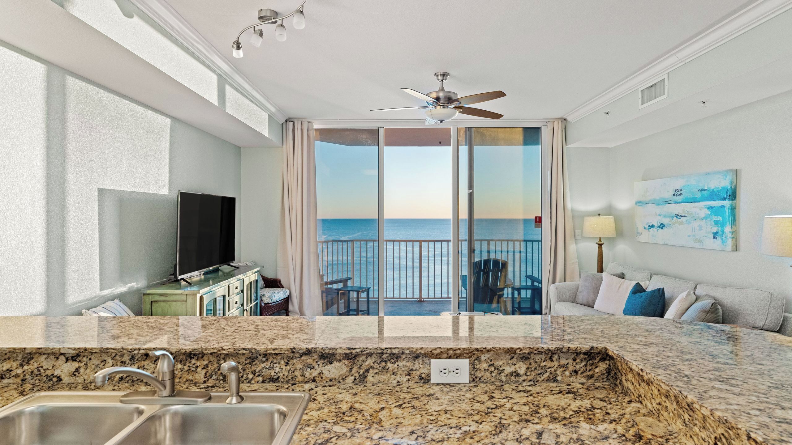 A modern hotel room with a kitchen area, a balcony with a view of the ocean, and a ceiling fan.