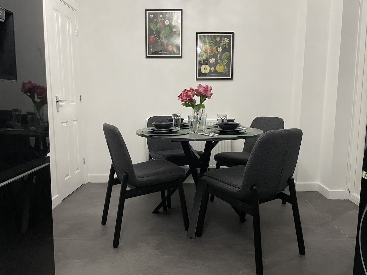 Een eetruimte met een ronde tafel, vier stoelen en een vaas met bloemen. Twee ingekaderde botanische prints sieren de muur.