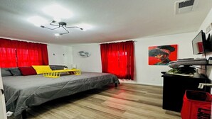 Une chambre à coucher avec un lit, des rideaux rouges, un ventilateur de plafond, un téléviseur fixé au mur et une petite table noire sur laquelle se trouve un sac rouge.