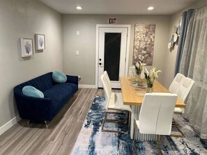 Una habitación moderna con un sofá azul oscuro, una mesa de comedor de madera con sillas blancas y una alfombra estampada en azul.