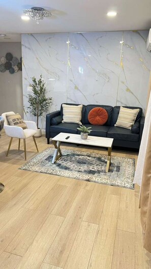 A modern living room with a dark blue sofa, a white armchair, a coffee table, and a marble-patterned wall. - Gillespie Dr 3BR 2BA Unit B | Near Downtown SD (Spring Valley)