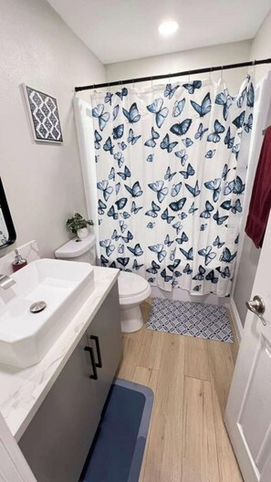 A bathroom with a shower curtain featuring a butterfly pattern, a white sink, a toilet, and a small potted plant. - Gillespie Dr 3BR 2BA Unit B | Near Downtown SD (Spring Valley)