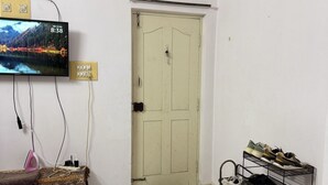 A room with a television mounted on the wall displaying a scenic image, a door with a doorknob, a shoe rack with multiple pairs of shoes, and a bed with a patterned blanket. - Fantastic 1-bedroom hall & Kitchen apartment in vibrant Bengaluru for your stay (Bengaluru)
