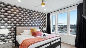 Quarto moderno com papel de parede estampado, uma janela grande com vista, uma cama com almofadas e uma mesa de cabeceira com abajur.