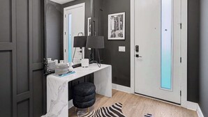 Um corredor com parede cinza escura, porta branca com painel de vidro fosco, mesa consola branca com tampos de mármore, cadeira preta e um tapete com padrão de zebra.