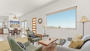 A spacious living room with a large window, a dining area, and a kitchen in the background. - Juniper House - Desert Retreat near JTNP - Stargazing, Views & Spa (Joshua Tree)