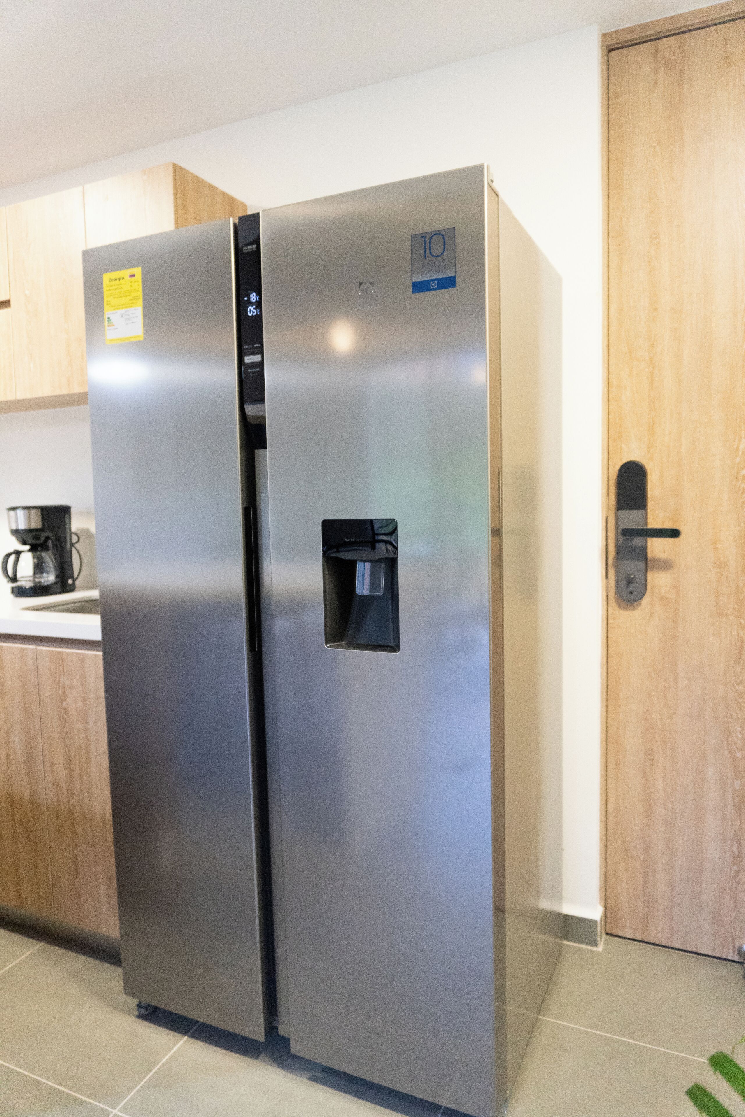 Un refrigerador de acero inoxidable con dispensador de agua, una cafetera sobre la encimera y una puerta de madera.