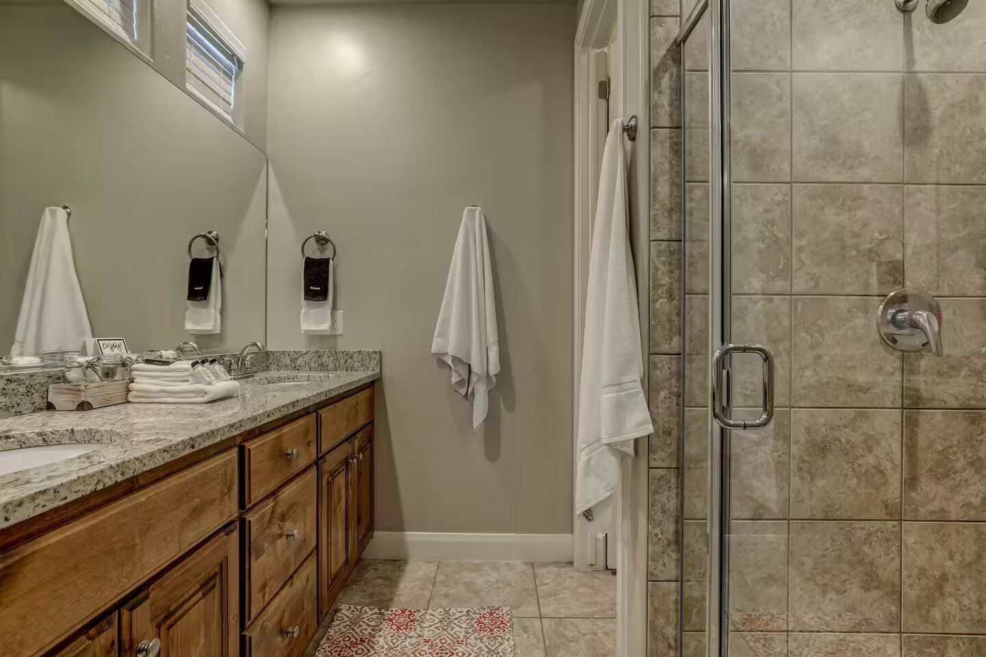 A bathroom with a granite countertop, wooden cabinets, a glass shower enclosure, and a patterned rug.