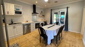 A modern kitchen with white cabinets, a stainless steel dishwasher, and a black range hood. There is a dining table with chairs and a window with curtains. - Central 1940s Bendigo Home Hamptons Heritage Reno (Long Gully)