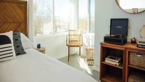 Et soveværelse med seng, stol, lille bord, pladespiller og et vindue med udsigt til træer.