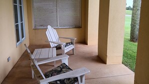 A covered patio area with two white chairs and a table, a window with blinds, and a view of a green lawn. - Villa C-Villa Ensuite (Gulfshores)