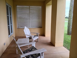 Un porche cubierto con dos sillas Adirondack blancas y una mesa, una ventana con persianas, y vista a un césped verde.