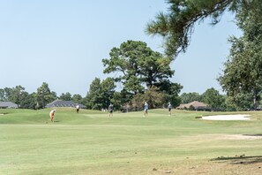 Un campo de golf bajo un cielo despejado, con un árbol grande y varias personas jugando.