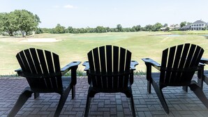 Three black Adirondack chairs on a brick patio overlooking a golf course with a water hazard and a clubhouse in the distance. - Villa D-Villa Ensuite (Gulfshores)