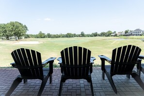 Tres sillas Adirondack negras sobre un patio de ladrillo con vistas a un campo de golf, un obstáculo acuático y un club de campo a lo lejos.