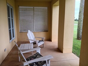 Een overdekt terras met twee witte stoelen en een tafel, een raam met zonwering, en uitzicht op een groen gazon.