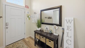 Un pasillo con una puerta blanca, una señal de salida, una consola con cestas, un espejo y un letrero que dice 'WELCOME'.
