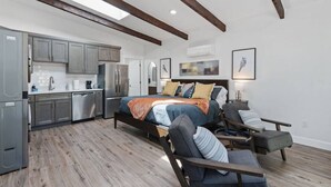 A modern bedroom with a large bed, a sitting area with a chair, and a kitchen area with cabinets and appliances. - Historic 1907 SLC Victorian - Hot Tub - Firepits - (South Salt Lake City)