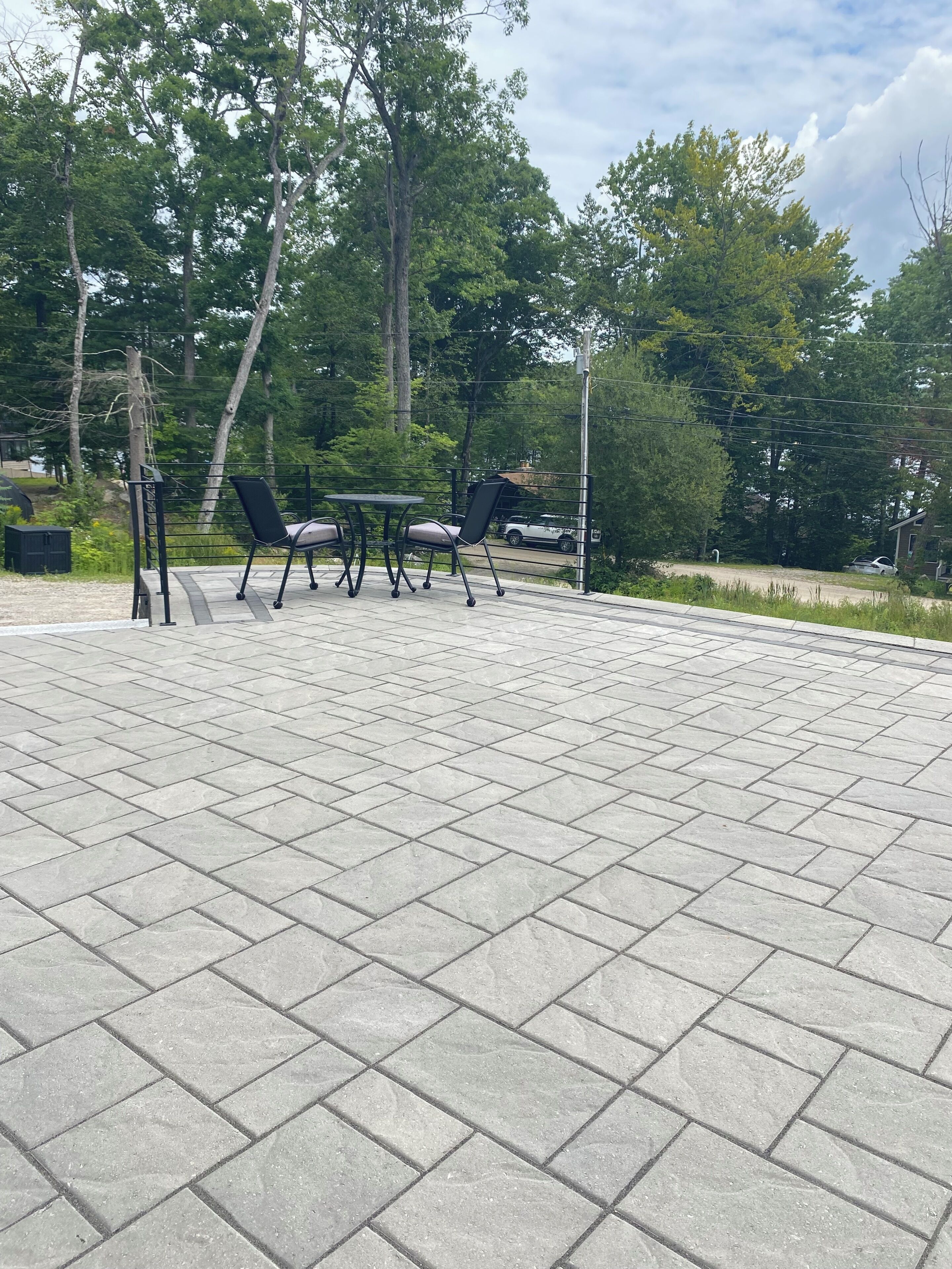 A paved patio area with a table and chairs, surrounded by trees.