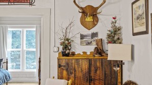 A room with a wooden cabinet, a deer head mounted on the wall, a small Christmas tree, and a window with curtains. - Camp Red Lodge - Cozy & Convenient (Red Lodge)