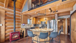 A log cabin interior with a central island, bar stools, a wood-burning stove, a kitchen with appliances, and a loft area. - Henry's Hideaway Cabin (Red Lodge)